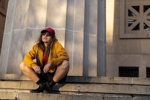 Latin American woman curly hair red cap sunglasses sitting on stairs with relaxed attitude