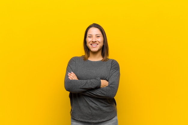 Latin american woman looking like a happy, proud and satisfied achiever smiling with arms crossed isolated against yellow wall