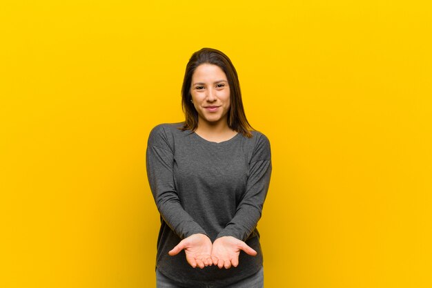 Latin american woman smiling happily with friendly, confident, positive look, offering and showing an object or concept isolated against yellow wall