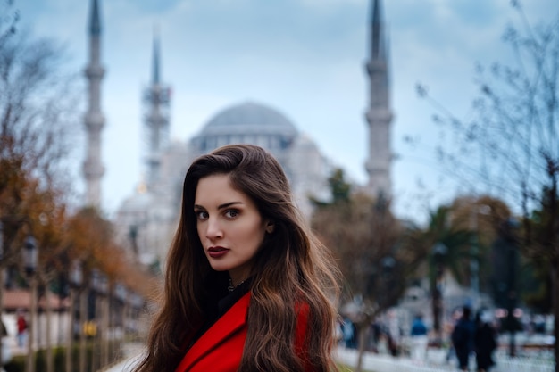 Photo latin american woman or turkish woman in a red stylish coat in front of the famous blue mosque in istanbul, photo of traveler on the background of a mosque on autumn day.