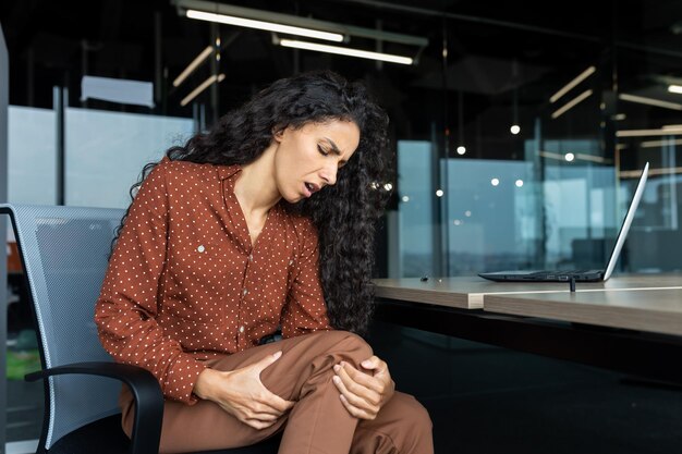 Latin american woman working inside office business woman has severe leg pain massaging muscle while