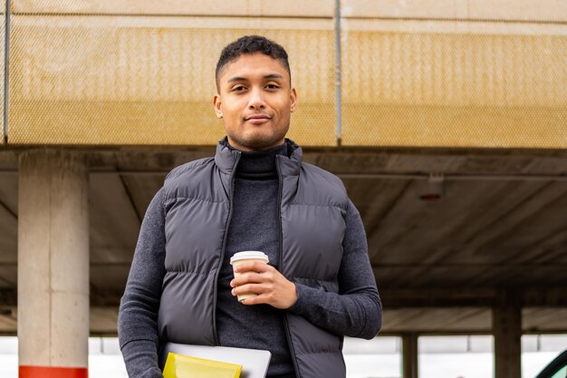 Latin businessman in the street holding laptop and a coffee to go Young latino man in jacket holding laptop and a coffee in the street