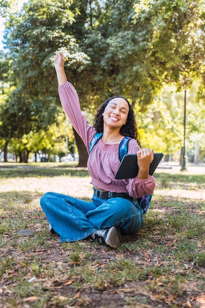 Latin college student woman celebrating and holding a tablet sitting outside in a park on the grass