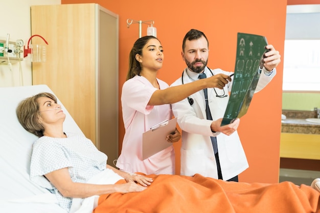 Latin doctors discussing over xray diagnosis while senior patient lying on bed in hospital