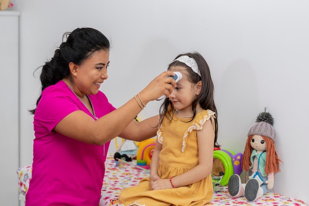 Latin female pediatrician checking the temperature of a girl with a dress sitting on the stretcher in the office