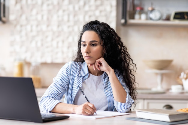 Latin lady studying remotely from home looking at laptop and making notes sitting at table in