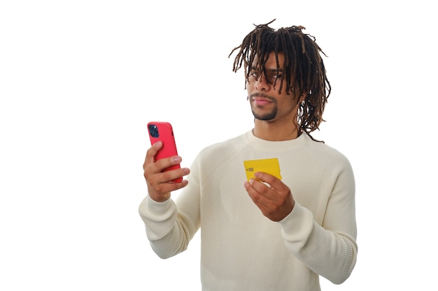 Latin man holding a mobile phone and a credit card while standing over an isolated background.