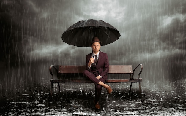 Latin man sitting on a bench with umbrella in the rain in the middle of a stormy night, horizontal