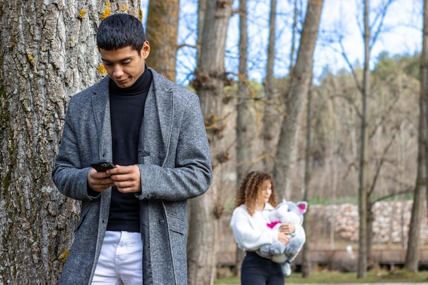 Latin man texting behind a tree while girlfriend approaches his with a teddy bear