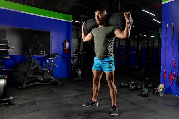 Latin man with sportswear carrying a sandbag on his back while training in a gym