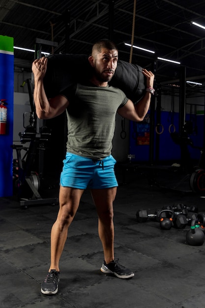 Latin man with sportswear carrying a sandbag on his back while training in a gym