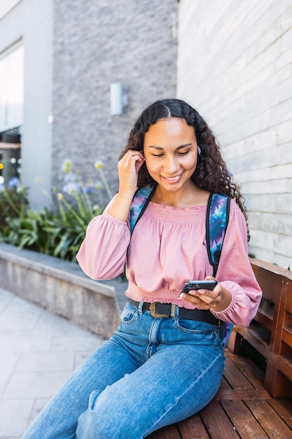 Latin university student woman smiling and using her mobile sitting outside the mall in free time