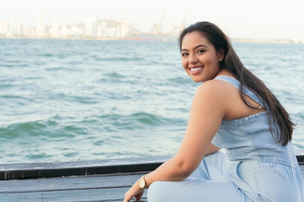 Latin woman is sitting on a pier smiling on a summer day