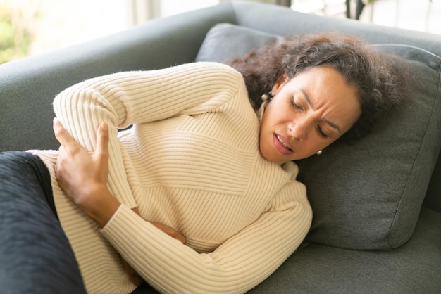 Latin woman lying down on sofa with stomachache feeling