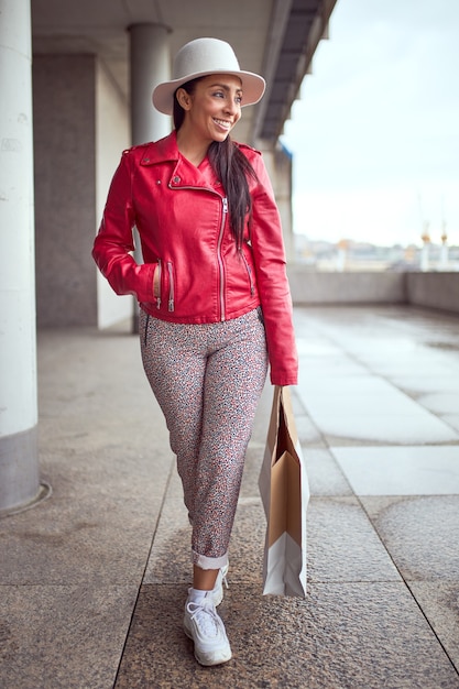 Latin woman in red jacket and white hat smiling with shopping bag.