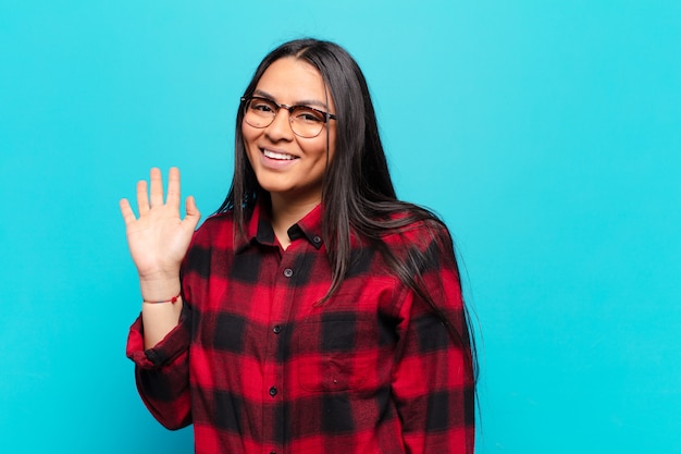 Latin woman smiling happily and cheerfully, waving hand, welcoming and greeting you, or saying goodbye