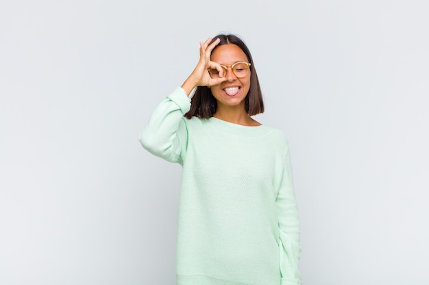 Latin woman smiling happily with funny face, joking and looking through peephole, spying on secrets