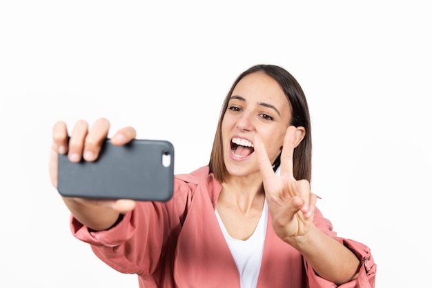 Latin young woman taking a selfie while making the victory sign over white background.