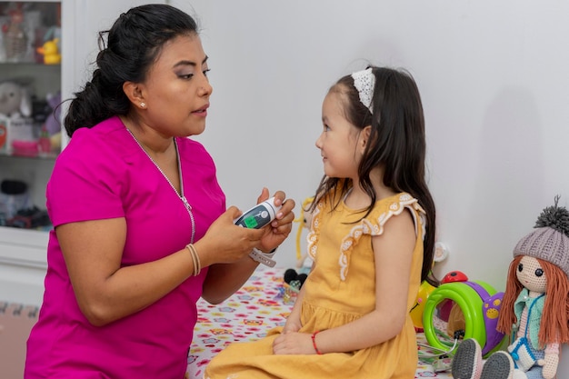 Latina pediatrician woman with a thermometer about to check the temperature of a girl sitting on the table in her office