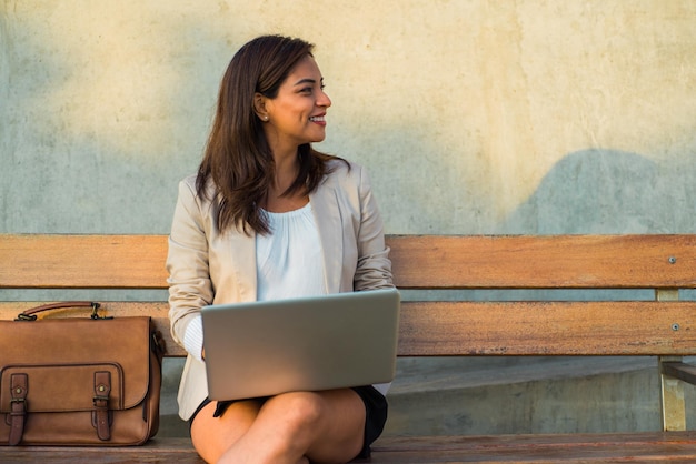 A Latina woman sitting on a bench with her computer looks smiling at the copy space