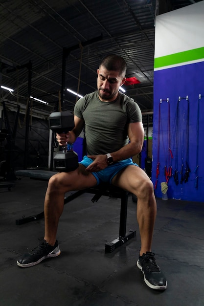 Latino man in sportswear holds a dumbbell on his legs while training in the gym