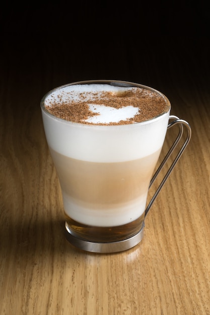 Latte art coffee with syrup, milk and cinnamon in a transparent glass with a metal handle on a wooden table