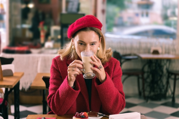 Latte in cafeteria. Elegant French woman wearing nice earrings and red coat drinking latte in cafeteria