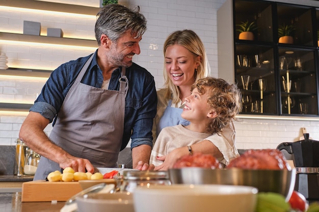 Laughing adult man in blue shirt showing son how to cook while woman standing near and cuddling boy