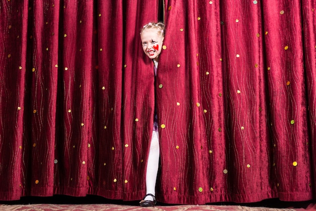 Laughing attractive young blond girl in colorful makeup making her entry on stage during a play or pantomime stepping through the closed curtains