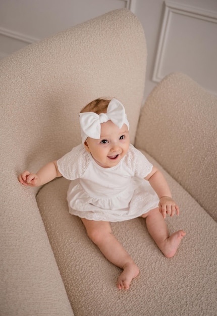 A laughing baby girl in a white dress and a bandage sits on a chair in the room and looks at the camera