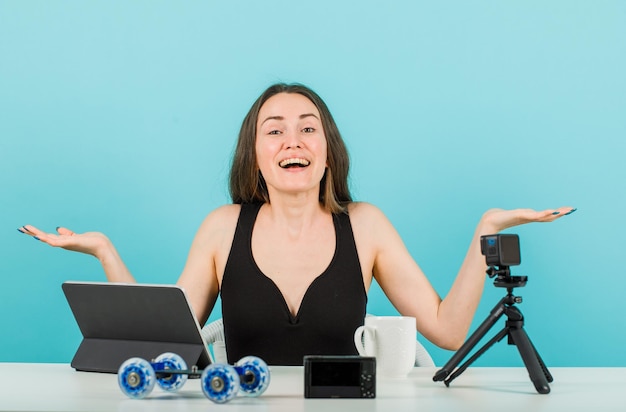 Laughing blogger girl is looking at camera by opening wide her hands on blue background