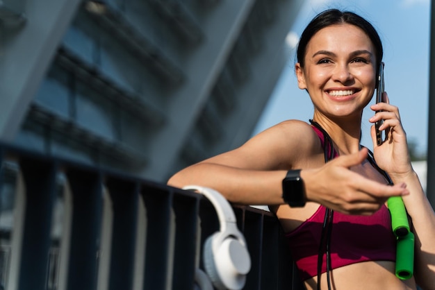 Laughing brunette woman with rejoiced face smiling wide and using mobile phone while chatting with somebody at the street after workout