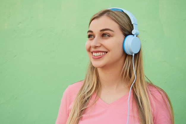 Laughing female student in a pink tshirt listening to music in blue headphones while standing against a green wall