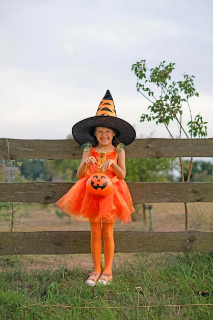 Photo a laughing girl dressed up in a halloween costume and hat holds bucket in the shape of a pumpkin