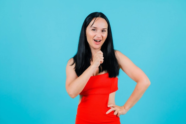 Laughing girl is holding her fist near chin and putting other hand on waist on blue background