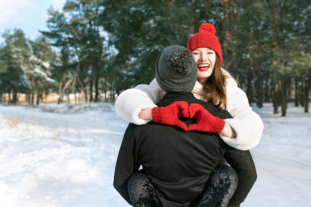 Laughing girl keeps hands in heart shape sign sitting on her guy hands Young loving couple in winter forest Sunny day