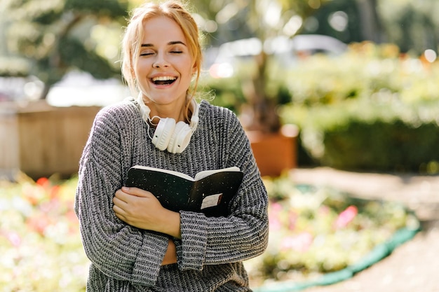 Laughing girl in knitted sweater holding textbook Outdoor portrait of adorable female student in headphones chilling in sunny day