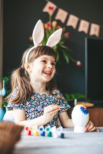 Laughing happy little girl with bunny ears painting Easter egg at home