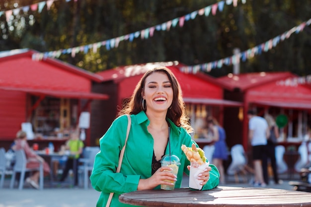 Laughing happy woman in green shirt spending time outdoors at fair in summer holding fast food