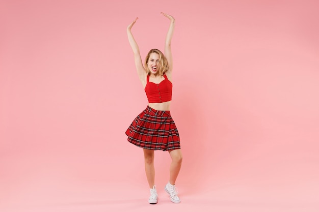 Laughing joyful young blonde woman girl in red sexy clothes posing isolated on pastel pink background studio portrait. People sincere emotions lifestyle concept. Mock up copy space. Rising hands up.