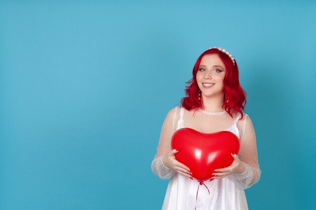 Laughing joyful young woman in a white dress with red hair holds a heart-shaped flying balloon