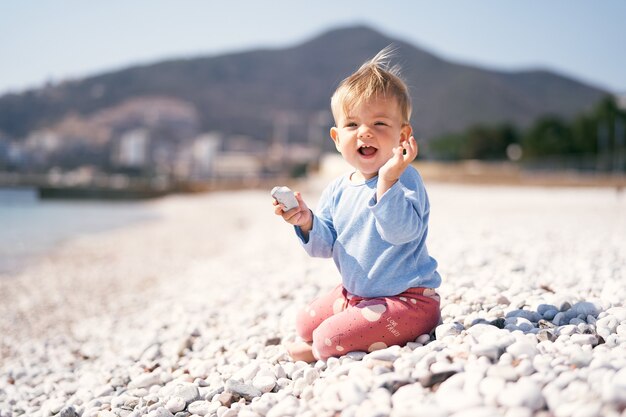 Laughing kid sits with a pebble in one hand and a raised second hand on a pebble beach