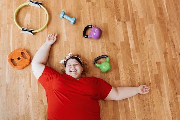 Laughing overweight woman dressed in sportswear and with a bandage on her head is laying on the floor next to the sports equipment