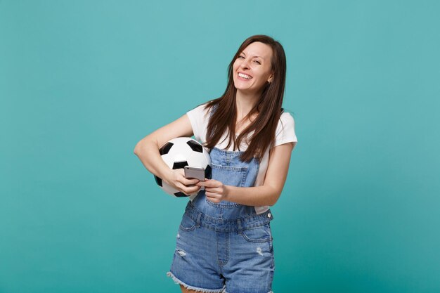 Laughing pretty young woman football fan holding soccer ball using mobile phone isolated on blue turquoise wall background. People emotions sport family leisure lifestyle concept. Mock up copy space.