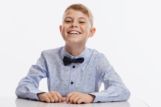 Laughing schoolboy in a blue shirt with a bow tie sits at the table. Back to school. White background.