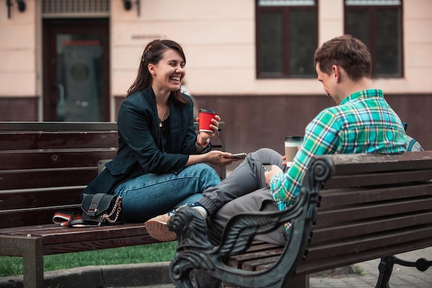 Laughing talking man with woman sitting on bench drinking coffee surfing in internet urban lifestyle