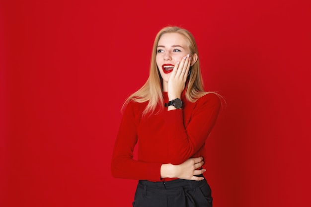 Laughing woman stands on a red background in casual clothes touching her face