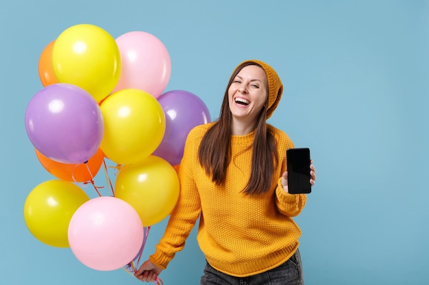 Laughing woman in sweater hat posing isolated on blue background. Birthday holiday party people emotion concept. Mock up copy space. Celebrating hold air balloons mobile phone with blank empty screen.