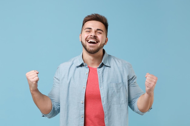 Laughing young bearded guy 20s in casual shirt posing isolated on pastel blue wall background studio portrait. People sincere emotions lifestyle concept. Mock up copy space. Doing winner gesture.