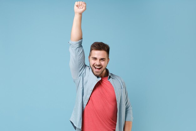 Laughing young bearded guy in casual shirt posing isolated on pastel blue background studio portrait. People sincere emotions lifestyle concept. Mock up copy space. Rising hand up, clenching fist.